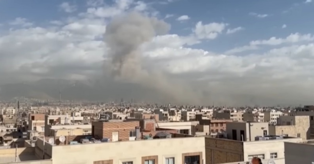 Vista aérea de uma área urbana residencial com prédios baixos e casas, onde uma grande coluna de fumaça cinza escura sobe ao céu após uma explosão. - Foto: Reprodução/Youtube