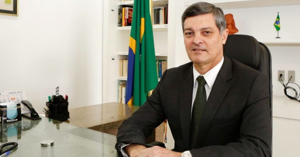Desembargador sentado em gabinete institucional, usando terno e gravata, com bandeira do Brasil ao fundo.