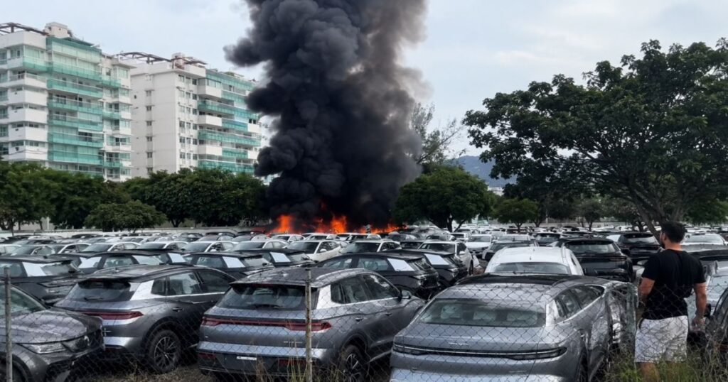 Incêndio de grandes proporções após queda de helicópteros no Recreio dos Bandeirantes, no Rio de Janeiro, com fumaça preta sobre estacionamento de veículos.
