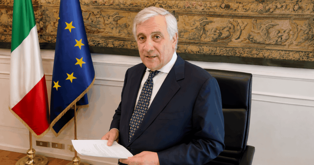 Autoridade italiana em escritório oficial segurando documento, com bandeiras da Itália e da União Europeia ao fundo.