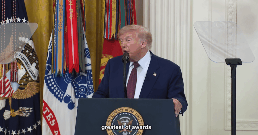 Donald Trump discursando em um púlpito durante cerimônia oficial na Casa Branca, com bandeiras militares e selo presidencial ao fundo.