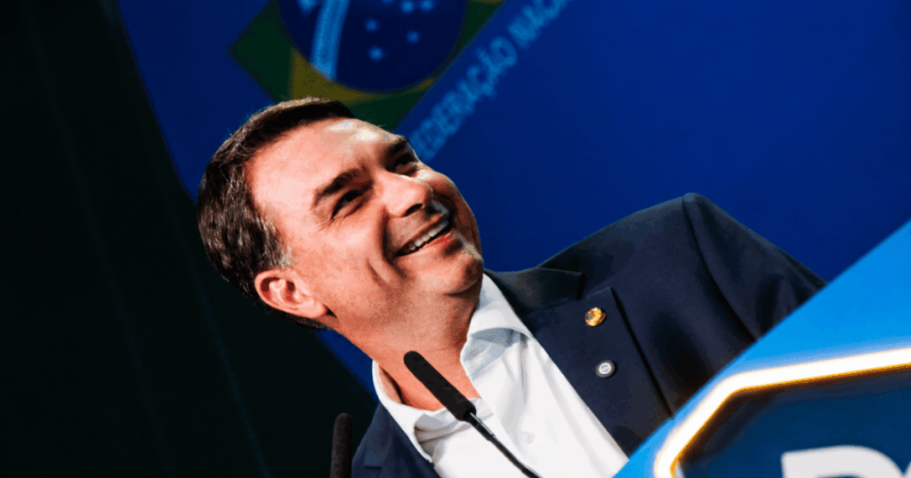 Homem sorrindo durante discurso em evento institucional, falando em púlpito com bandeira do Brasil ao fundo.