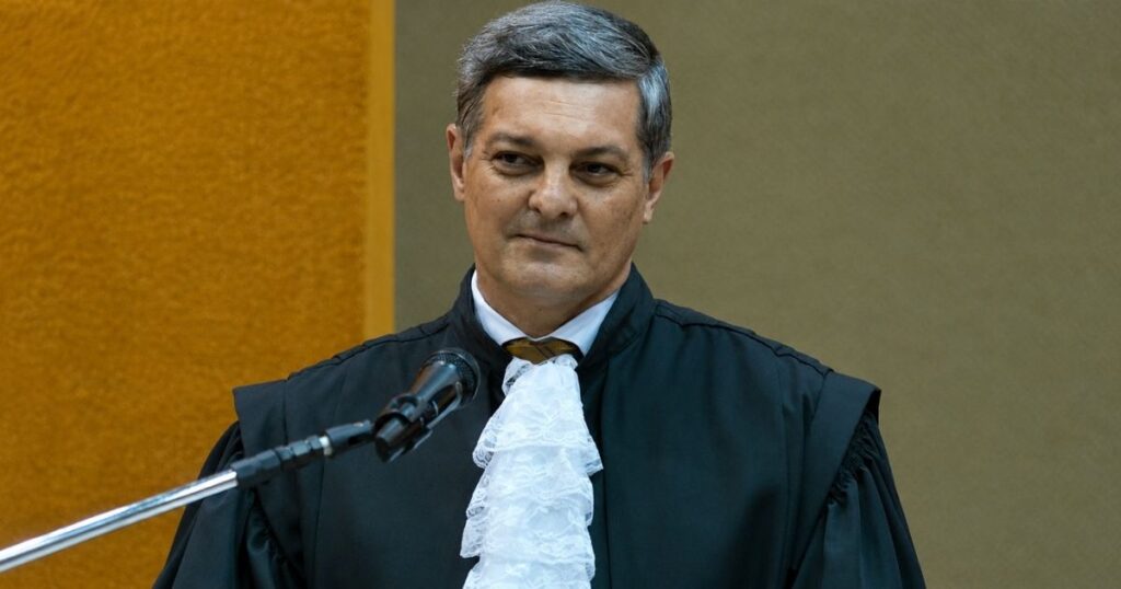 Desembargador trajando veste oficial durante cerimônia institucional, posicionado diante de um microfone em ambiente formal.