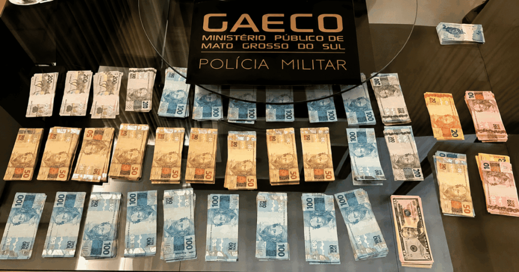 Mesa com cédulas de real e um painel do GAECO do Ministério Público de Mato Grosso do Sul e da Polícia Militar durante apresentação de dinheiro apreendido em operação.