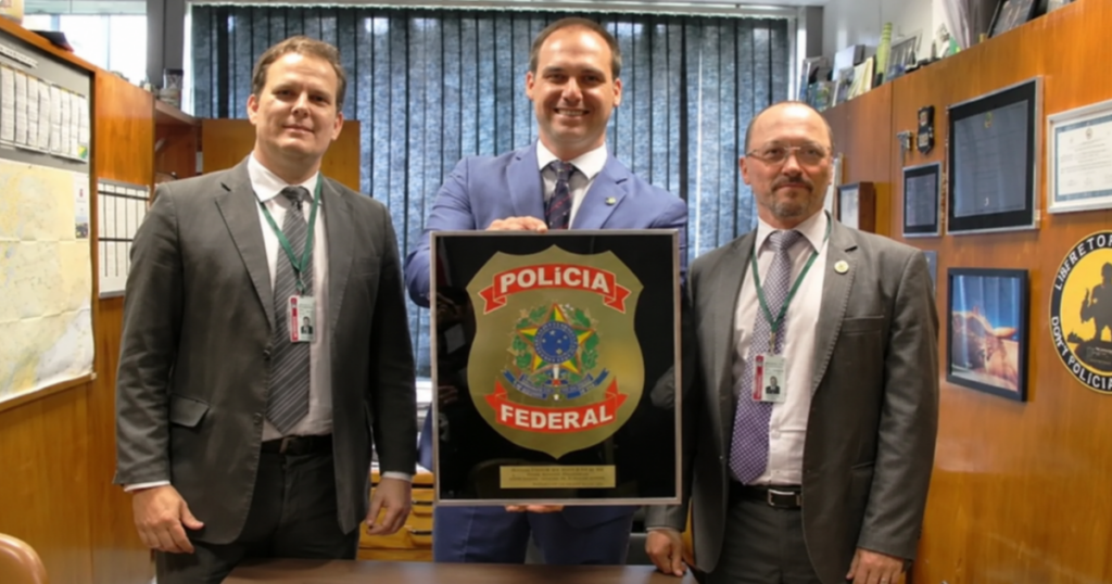 Eduardo Bolsonaro segura quadro com símbolo da Polícia Federal ao lado de dois homens em escritório