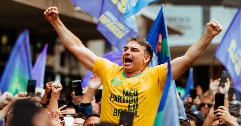 Flávio Bolsonaro ergue os braços durante ato de campanha com apoiadores e bandeiras em Juiz de Fora.
