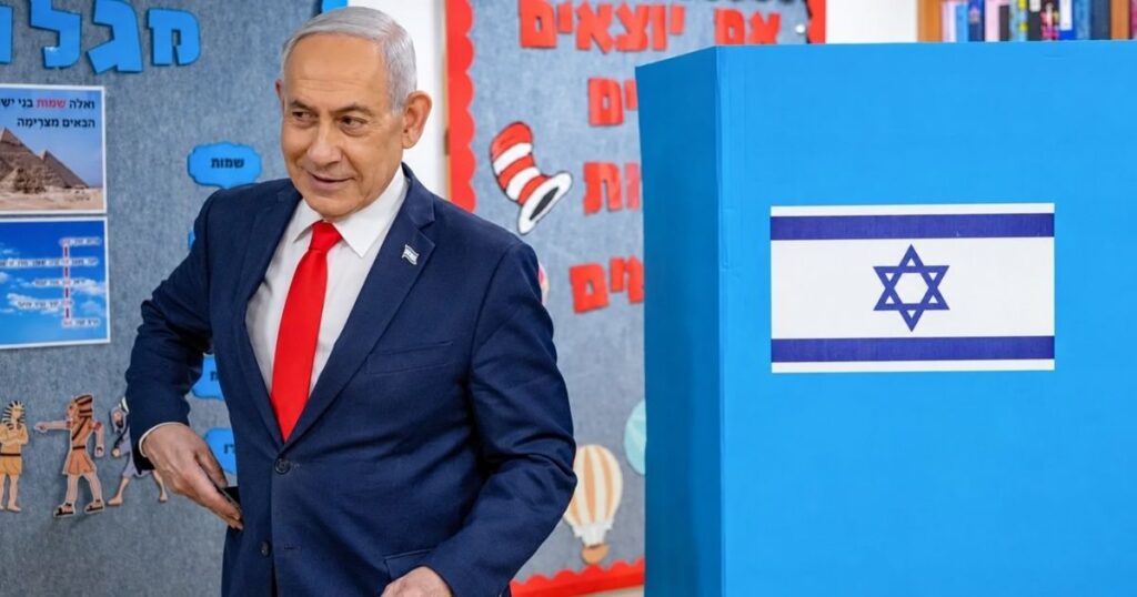 Benjamin Netanyahu, primeiro-ministro israelense, aparece ao lado de uma cabine de votação com a bandeira de Israel, em Jerusalém, durante as eleições de 27 de outubro de 2026.