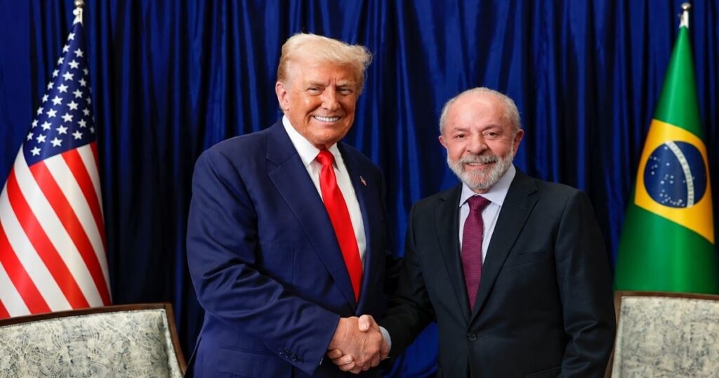 Donald Trump e Lula apertam as mãos durante encontro oficial, com as bandeiras dos Estados Unidos e do Brasil ao fundo.