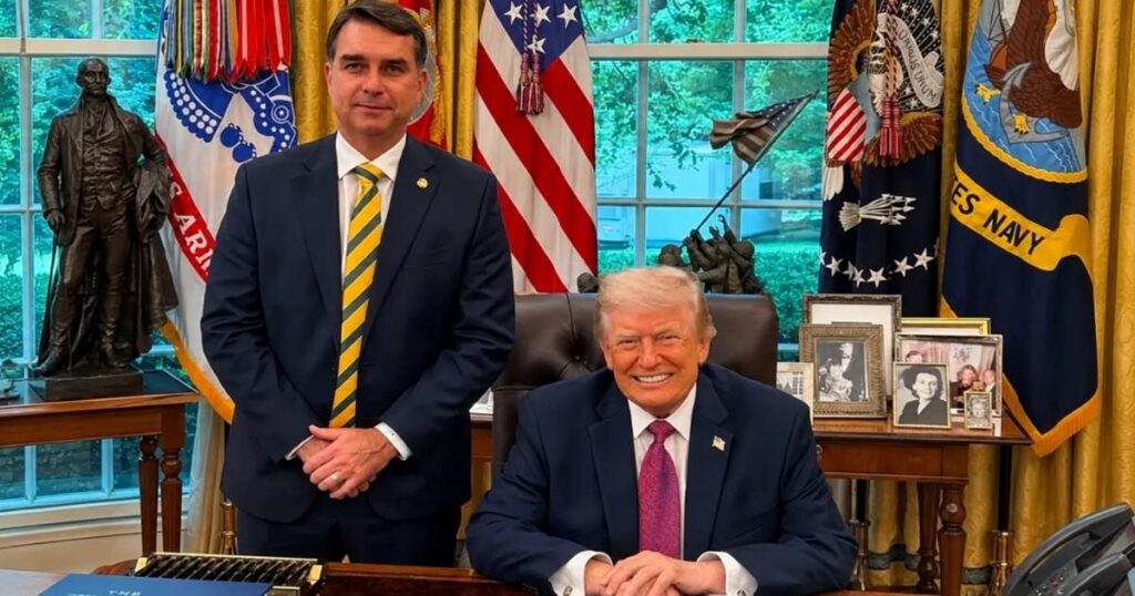 Donald Trump aparece sentado à mesa no Salão Oval da Casa Branca, ao lado do senador Flávio Bolsonaro, que está em pé, com bandeiras dos Estados Unidos ao fundo.