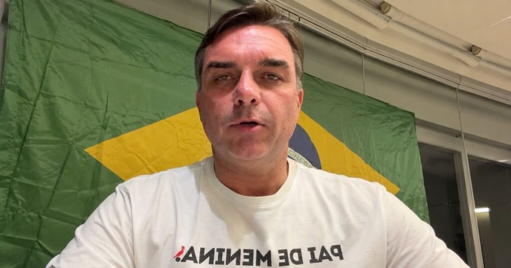 Flávio Bolsonaro aparece diante da bandeira do Brasil, usando camiseta branca, em imagem que ilustra matéria sobre possível mudança para os EUA após derrota para Lula.
