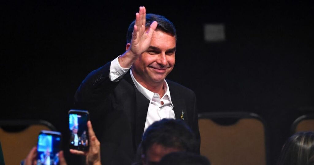 Flávio Bolsonaro sorri e acena ao público durante evento. Candidato propõe manter o Bolsa Família por até dois anos após emprego formal.