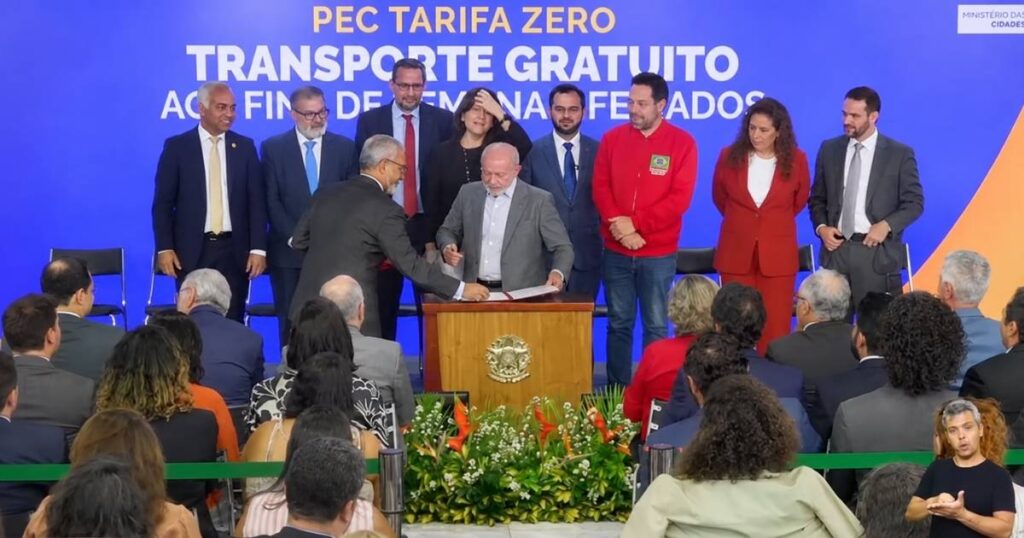 Presidente Lula aparece diante de documentos durante cerimônia oficial, acompanhado por autoridades, com público e intérprete de Libras.